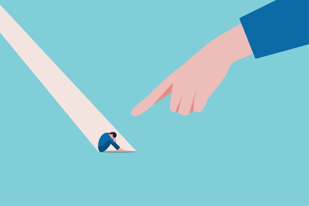 A large hand points at a small, sad person sitting at the end of its shadow on a blue background, symbolizing blame, criticism, or feeling pressured.