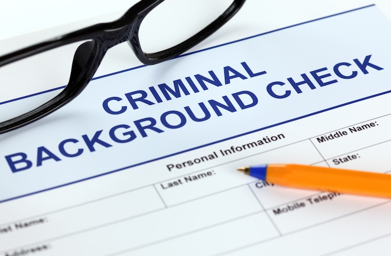 A close-up of a form labeled "Criminal Background Check" with sections for personal information. A pair of glasses and an orange pen rest on the form, highlighting the importance of Internet reputation monitoring during background checks.