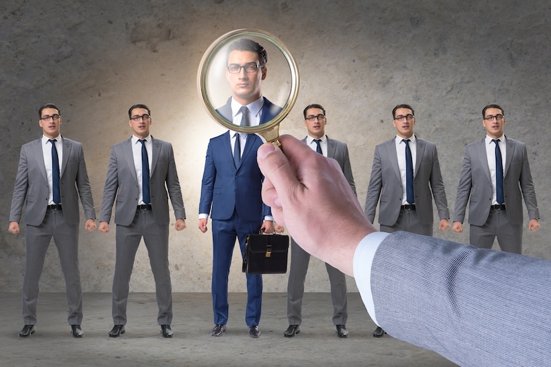A person in a suit holds a magnifying glass in front of one of five identical businessmen standing in a row, highlighting the face of the selected individual as part of a background check.