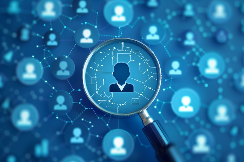 A magnifying glass hovers over a silhouette of a person among many connected user icons on a blue digital network background, symbolizing searching or analyzing people within a network without conducting a background check.