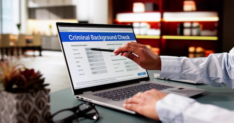 A person uses a laptop displaying a “Criminal Background Check” form, holding a pen and pointing at the screen. The scene appears to be in an office with bookshelves, possibly seeking ways to remove background check records.