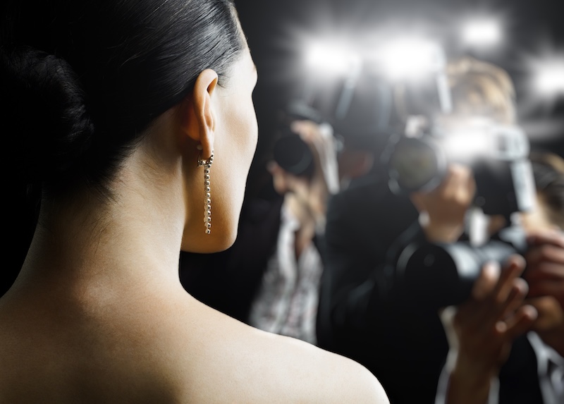 A woman with an elegant updo and dangling earring faces away from the camera as paparazzi, chasing a revenge porn scandal, take photos of her, bright flashes illuminating the scene.