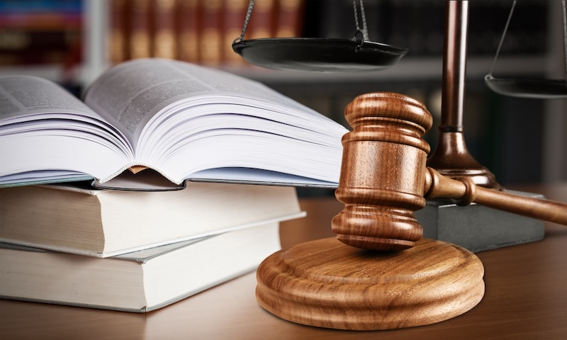 A wooden judge’s gavel rests on a sound block beside two stacked books and an open book, with scales of justice and law books blurred in the background, symbolizing legal action against revenge porn.