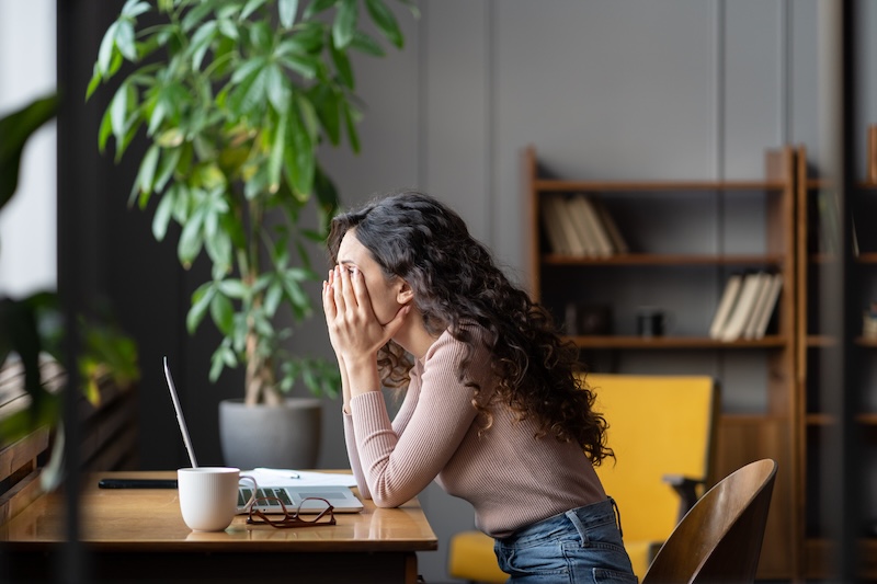 A woman with long curly hair sits at a desk with her face in her hands, appearing stressed—possibly overwhelmed by issues like revenge porn. A laptop, notebook, and coffee mug are on the desk. Shelves with books and a large plant are in the background.