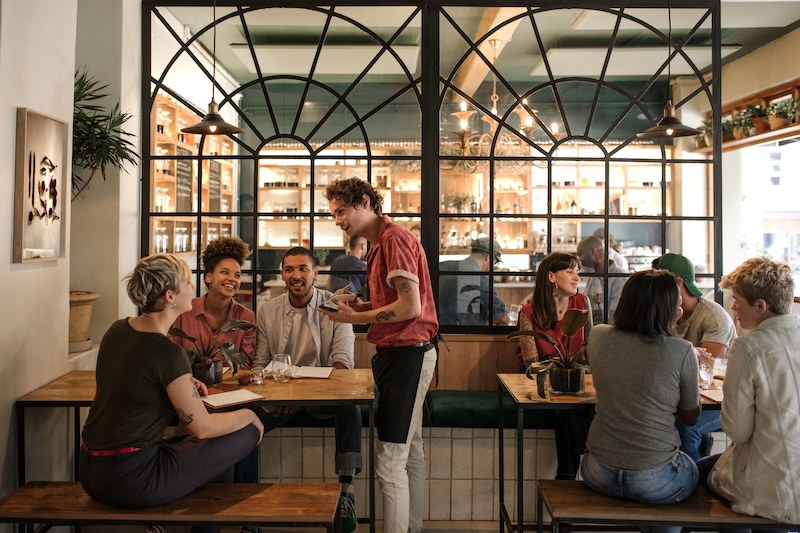 A server takes orders from a group of smiling customers at a modern, stylish restaurant—proof that the venue is working to recover from a damaged reputation, with diners seated nearby and shelves of bottles visible through a large window in the background.