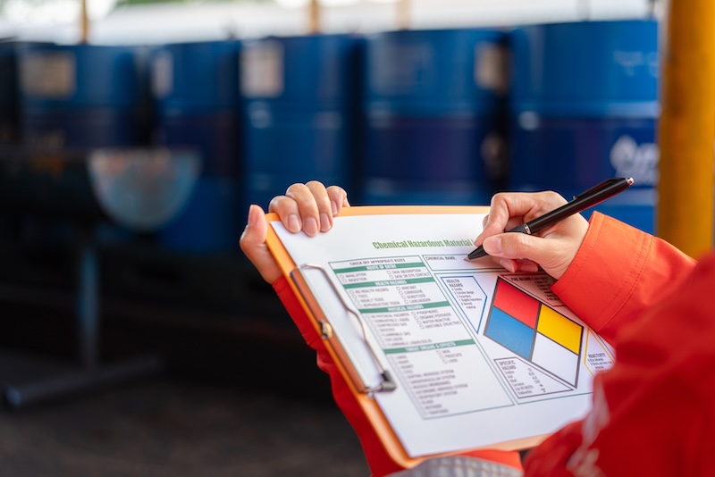A person wearing an orange safety jacket holds a clipboard and pen, checking a hazardous materials information sheet in front of several blue industrial barrels—an essential step to recover from a damaged reputation in safety compliance.