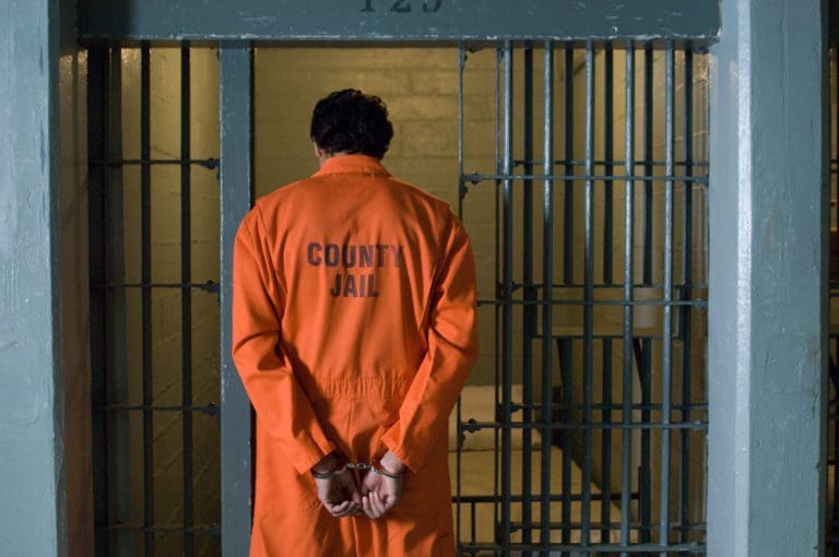 A man in an orange prison uniform standing in a jail cell.