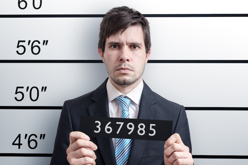 A man in a suit and tie stands against a height chart background, holding a sign with the number "367985." The chart shows height measurements in feet and inches. The man's neutral expression adds to the typical look often seen in mugshots, which are public record.