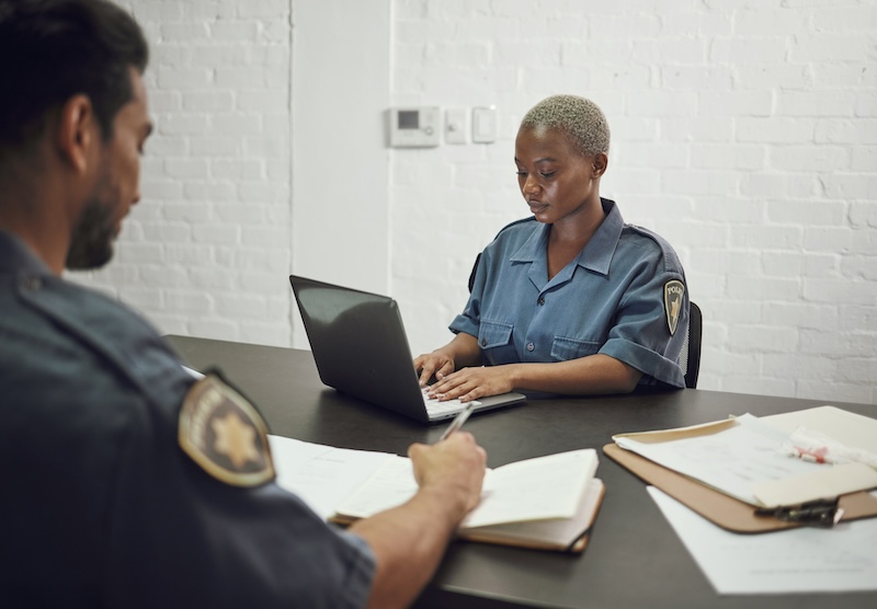 Two uniformed officers sit at a table with paperwork. The officer in the foreground is focused on writing, while the officer in the background is working on a laptop. Both appear to be in a serious discussion, possibly in an office or briefing room, perhaps debating whether mugshots are public record.