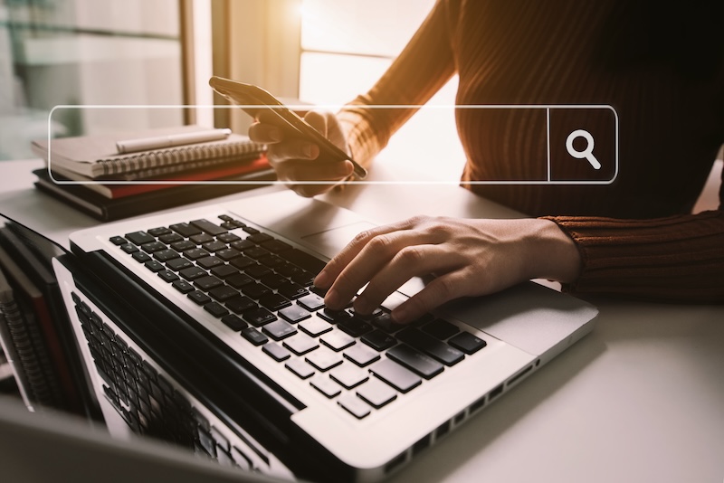 A person multitasks at a desk, using a smartphone and typing on a laptop, with a transparent search bar icon overlaid—suggesting they’re searching online topics like "Are Mugshots Public Record in California?.