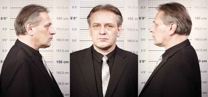 A middle-aged man in a suit is photographed in three different angles (left profile, front, and right profile) in front of a height measurement chart used for booking photos. The chart displays height in both centimeters and inches.