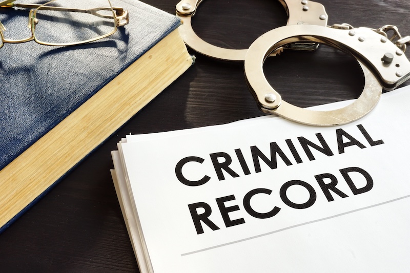 A pile of papers labeled "Criminal Record" lies on a dark surface next to a pair of handcuffs, a dark blue book, and a pair of eyeglasses.