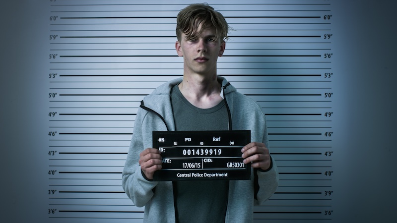 A young man wearing a gray hoodie and gray t-shirt stands in front of a height measurement chart holding a police identification sign. The chart shows measurements in feet and inches. The date on the sign reads 17/06/15 and it mentions Central Police Department.