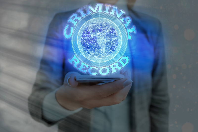 A person in a suit holding a smartphone with a holographic display above it. The display shows the words "CRIMINAL RECORD" encircling a digital globe, symbolizing the concept of accessing criminal records through technology.