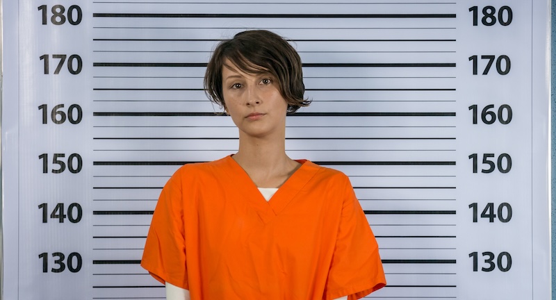 A person with short, dark hair stands in front of a height chart, wearing an orange jumpsuit. The height chart is marked with measurements ranging from 140 to 180 centimeters. The person is looking directly at the camera with a neutral expression.