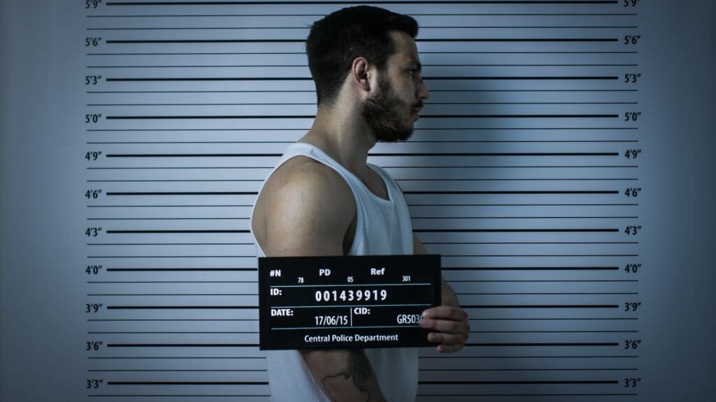 A man in a white tank top is standing in profile against a height chart holding a mugshot board with identification details. The board reads "Central Police Department" and "ID: 001439919" among other details.