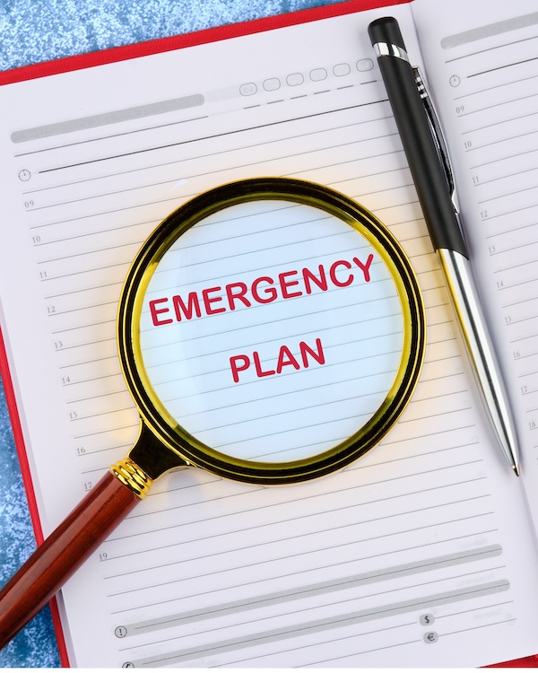 Magnifying glass focusing on the words "Emergency Plan" on an open book with blank pages, accompanied by a pen.