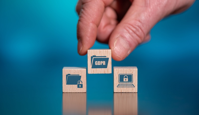 A hand arranges wooden blocks against a blue background. One block shows "GDPR" with a folder icon, another has a folder with a lock, and the third features a laptop with a lock, symbolizing data protection and privacy.