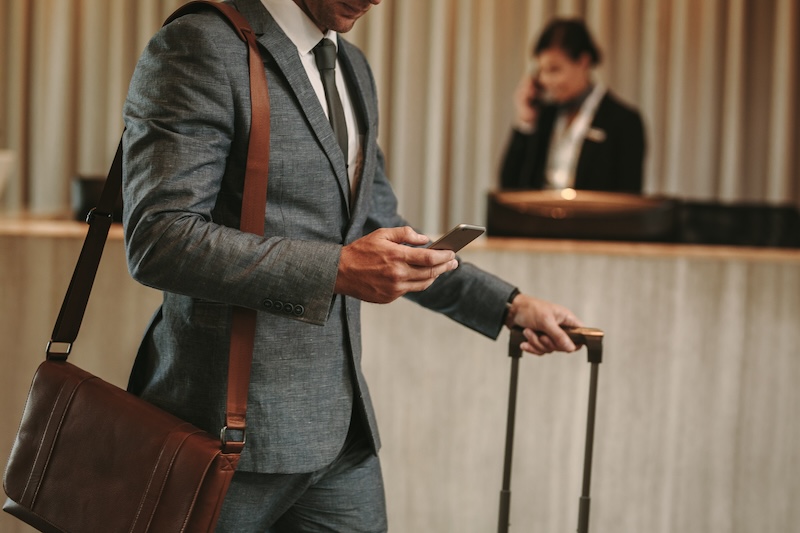 A man in a gray suit stands in a hotel lobby, holding a smartphone and a suitcase handle. A brown shoulder bag is slung across his body. In the background, a receptionist is on the phone at the front desk.