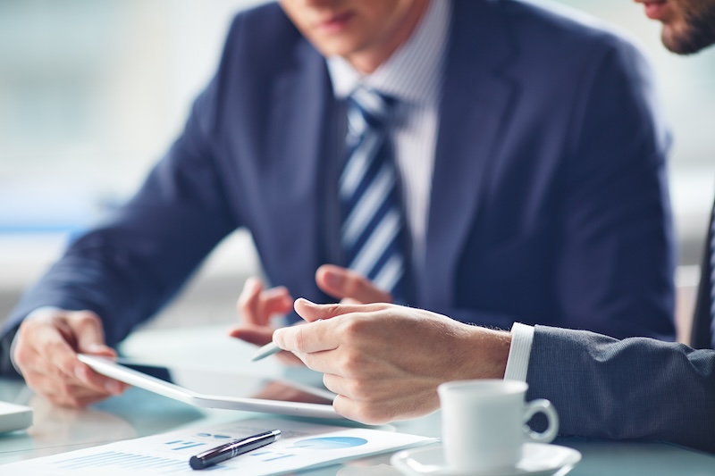 Two people in business attire are sitting at a table. One person holds a tablet, and they are discussing its contents. A document, pen, and coffee cup are on the table. The focus is on their hands and the tablet.