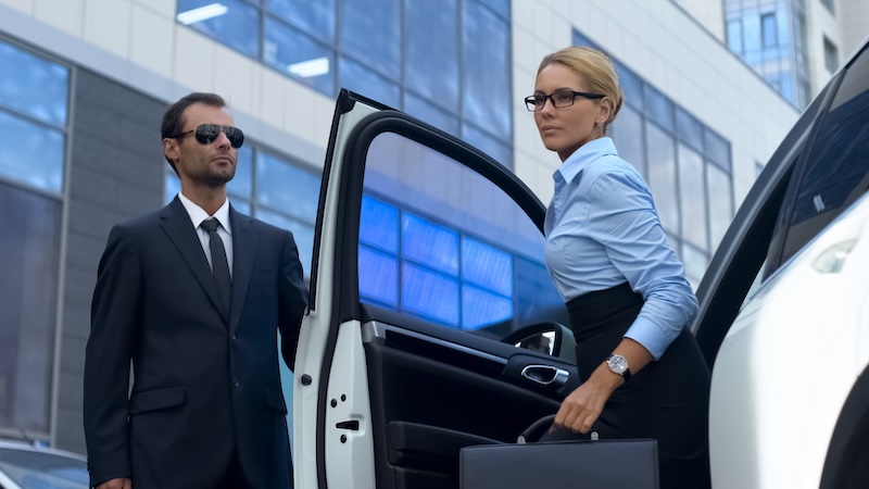 A woman in business attire and glasses exits a car, holding a briefcase. A man in a suit and sunglasses stands nearby. They are in an urban setting with modern buildings visible in the background.