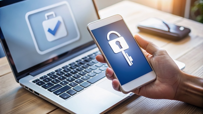 A person holds a smartphone displaying a lock and key graphic in front of a laptop screen with a locked briefcase icon and checkmark. A blurred smartphone lies on a table. The scene suggests themes of security and digital protection.