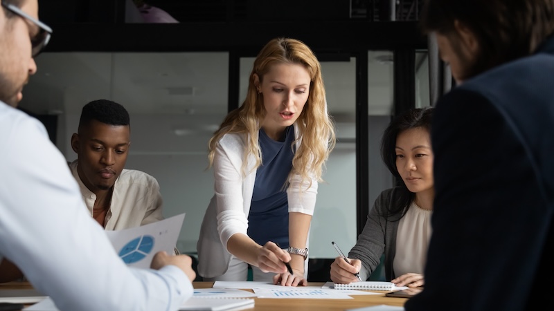 A diverse group of five people in a modern office setting engaged in a meeting. A woman stands and points to a document on the table as others listen and take notes. Charts and graphs are visible on some of the papers.