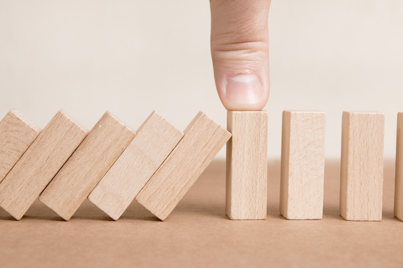 A row of wooden dominoes is falling, but a finger presses against one upright domino, stopping the chain reaction on a brown surface.