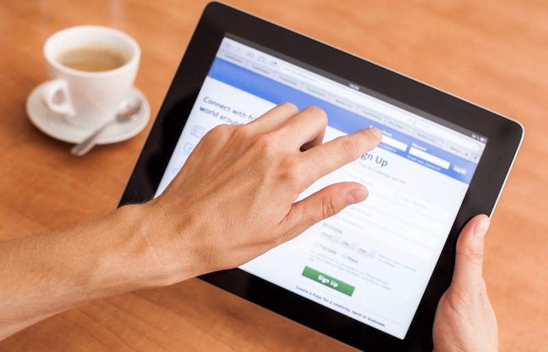 A person uses a tablet to browse a social media website on a wooden table. A cup of coffee sits nearby.