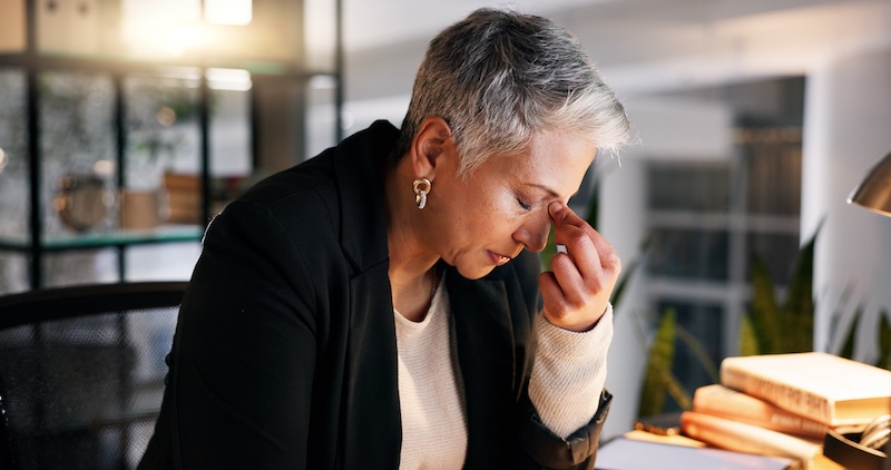 A person with short gray hair sits at a desk, pinching the bridge of their nose while looking down, appearing stressed or tired. They are indoors, surrounded by shelves, books, and a lamp.