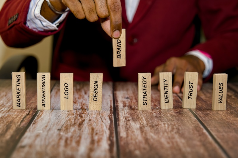 A person in a red suit arranges wooden blocks on a table. The blocks are labeled with words like "MARKETING," "ADVERTISING," "LOGO," "DESIGN," "BRAND," "STRATEGY," "IDENTITY," "TRUST," and "VALUES.