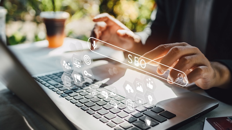 Person typing on a laptop outdoors with sunlight. Transparent floating icons related to SEO and digital marketing surround the keyboard, including a search bar with "SEO" highlighted, graphic charts, and social media symbols.