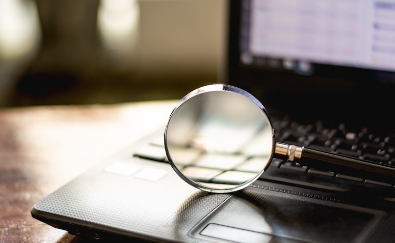 A magnifying glass rests on the keyboard of an open laptop, with a blurry screen displaying an application window. Sunlight illuminates the scene from the left, creating a focused reflection on the lens.