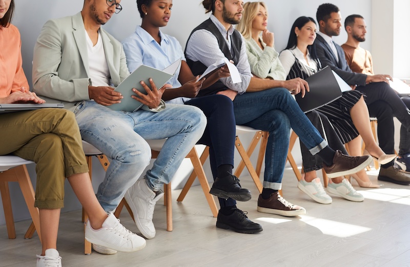 A diverse group of people sit in a row on chairs, holding resumes or folders, waiting for an interview in a bright room, their focused expressions revealing concern over overcoming a damaged reputation.